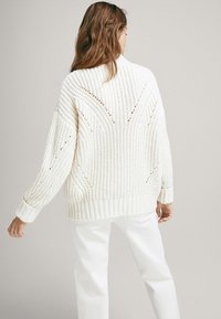 Nainen, joka on pukeutunut teksturoituun, väljävalkoiseen neulepuseroon ja valkoisiin housuihin, seisoo selkä katsojaan päin tasaisella taustalla.