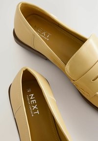 Geel leren loafers met een gladde afwerking, voorzien van een decoratief snijontwerp en een bruine rubberen zool. Branding zichtbaar aan de binnenzijde.