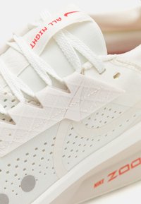 Nahaufnahme eines weißen Nike Zoom Laufschuhs mit perforiertem Mesh, weißen Schnürsenkeln und rotem Schriftzug "ALL NIGHT" sowie "NIKE ZOOM".