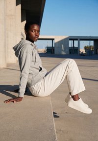 Mujer con sudadera gris con capucha, pantalones blancos y zapatillas blancas sentada en un bordillo de hormigón al aire libre bajo un cielo azul despejado.