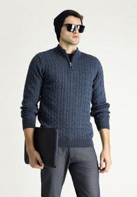 Marineblaue Strickpullover mit Reißverschlusskragen, kombiniert mit dunklen Jeans. Zum Zubehör gehört eine schwarze, strukturierte Laptoptasche und Sonnenbrille.