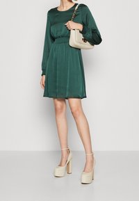 Grön satindress med långa ärmar, smockad midja och en svängig kjol. Bärs med beige högklackade skor med ankelrem och en krämfärgad axelväska.