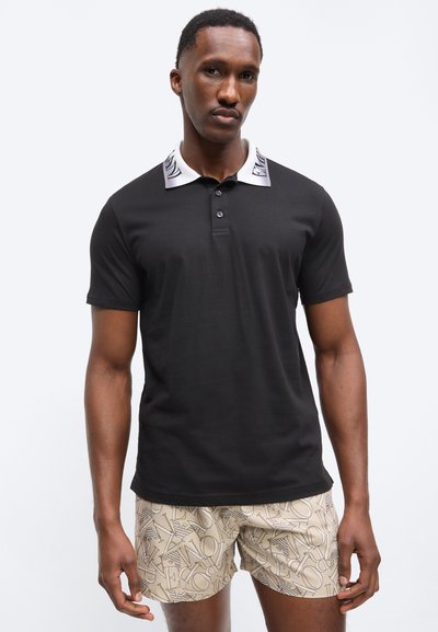Άνδρας που φοράει μαύρο πόλο μπλουζάκι με κοντά μανίκια και λευκό γιακά με σχέδιο, καθώς και μπεζ σορτς με αφηρημένο γράμμα τύπωμα.