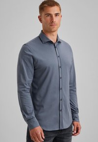 Joven con el cabello corto, vestido con una camisa gris de manga larga abotonada y jeans oscuros, de pie contra un fondo claro y liso.