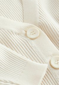 Cremefarvet strikket cardigan med ribbet tekstur, V-hals og store runde knapper. Blødt stof med synlige sømdetaljer.