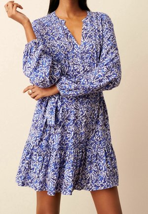 Kobieta ubrana w niebiesko-białą sukienkę w kwiaty z długimi rękawami, dekoltem w serek i falbanami, pozująca z założonymi skrzyżowanymi rękami.