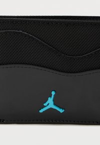 Crna novčanika s teksturiranim gornjim dijelom od tkanine i sjajnim crnim donjim dijelom. Ima istaknuti tealfarbanog Jumpman logotipa na prednjoj strani.