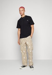 T-shirt nera in cotone con un piccolo logo, abbinata a pantaloni beige a fantasia con disegni floreali e astratti, completata da sneaker nere.