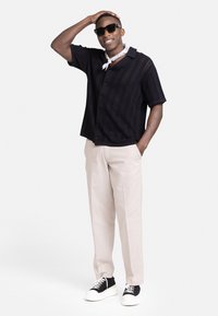 Camisa negra de botones con un patrón texturizado, combinada con pantalones beiges y zapatillas negras. El modelo lleva gafas de sol y un pañuelo blanco.