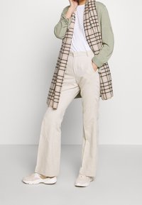 Un cardigan vert clair sur un t-shirt blanc, une écharpe à carreaux, un pantalon large beige et des baskets claires. Un ensemble texturé et décontracté aux tons neutres.