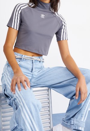 Personne assise portant un crop top gris Adidas avec des rayures blanches, un pantalon de survêtement Adidas bleu clair avec des rayures blanches et des baskets blanches.