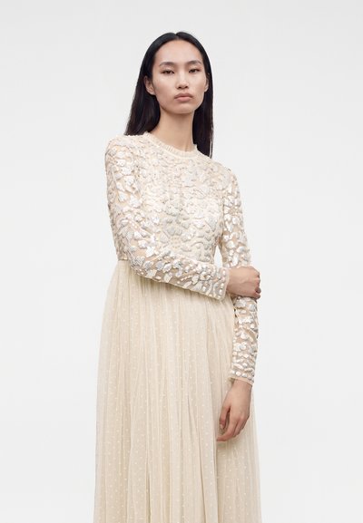 Femme aux longs cheveux foncés portant une robe crème à manches longues avec des sequins floraux sur le haut et une jupe plissée, debout devant un fond uni.