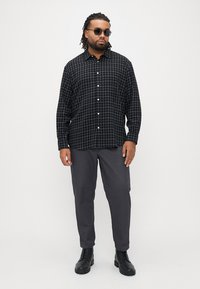 Camisa às quadrículas preta e verde feita de algodão, com design de abotoar, mangas compridas, usada com calças cargo cinzentas e botas pretas.