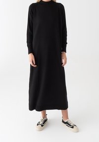 Osoba stojí v dlouhé černé svetr šatech v kombinaci s černobílými teniskami, na minimalistickém bílém pozadí.