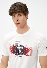 Camiseta blanca de algodón con un gráfico impreso en negro y rojo de una escena histórica de multitudes, con texto en turco y una firma cerca de la parte inferior.