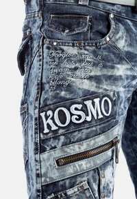 Pantaloni in denim in blu chiaro e scuro con un finish consumato. Presentano testo ricamato e la parola "KOSMO". Comprendono cerniere decorative e rivetti.