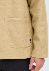 Beige wool blend jacket with a side pocket and a small black logo tag, featuring a simple and clean design with a soft texture.