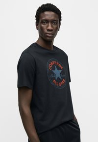 Μαύρο βαμβακερό t-shirt με κοντά μανίκια, με κυκλικό λογότυπο σε μπλε και κόκκινο, που γράφει "Converse All Star" και "Chuck Taylor."