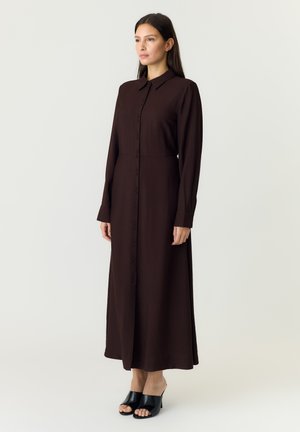 Femme debout portant une robe maxi marron foncé à manches longues, boutonnée, avec un col et des chaussures à talons ouvertes noires sur un fond clair.