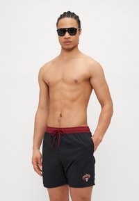 Calções de banho pretos com cordão vermelho, logótipo bordado e bolsos. O tecido parece leve e suave. O modelo usa óculos de sol.