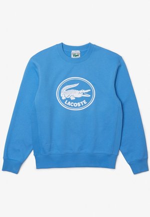 Niebieska bluza z okrągłym dekoltem i białym owalnym logo Lacoste z krokodylem i nazwą marki na środku.