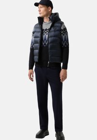 Navy puffer vest over een geblokt sweater, donkere broek en zwarte schoenen. Voorzien van een ritsluiting, capuchon en geometrische patronen op de sweater.