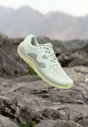 Zapatilla de trail running verde claro flotando sobre un terreno rocoso con un fondo montañoso desenfocado.