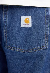 Denim jeans with a back pocket and blue fabric. Features a white label with an orange logo and text "carhartt" stitched in red.