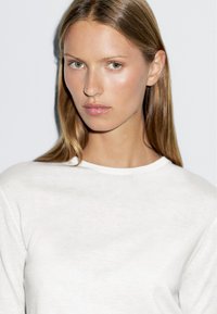 Witte gebreide trui met een ronde hals en lange mouwen, met een soepele textuur. Het model heeft steil haar en neutrale make-up.