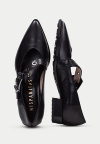 Zapatos de tacón de cuero negro con punta afilada. Cuentan con una correa en el tobillo con una hebilla de metal y un tacón cuadrado texturizado. Acabado suave.
