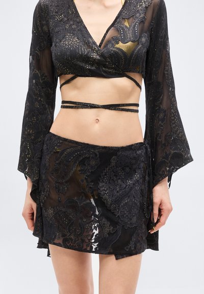 Ensemble crop top noir transparent et jupe portefeuille avec motifs cachemire dorés et fines bretelles autour de la taille, porté par une personne avec le ventre nu visible.