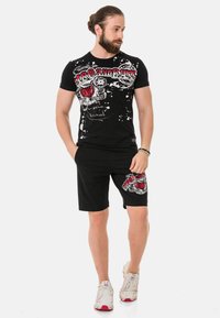 Schwarzes T-Shirt mit weißem und rotem Grafikdruck sowie Splattersymbolik, kombiniert mit schwarzen Shorts, die ähnliche Grafiken aufweisen. Lässige Sportbekleidung.