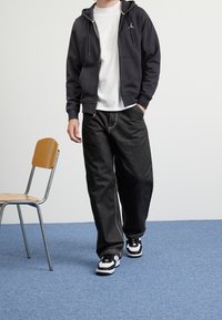 Persona che indossa una felpa nera con zip, una maglietta bianca, pantaloni neri larghi, e sneakers bianco e nere, in piedi vicino a una sedia di legno su un tappeto blu.