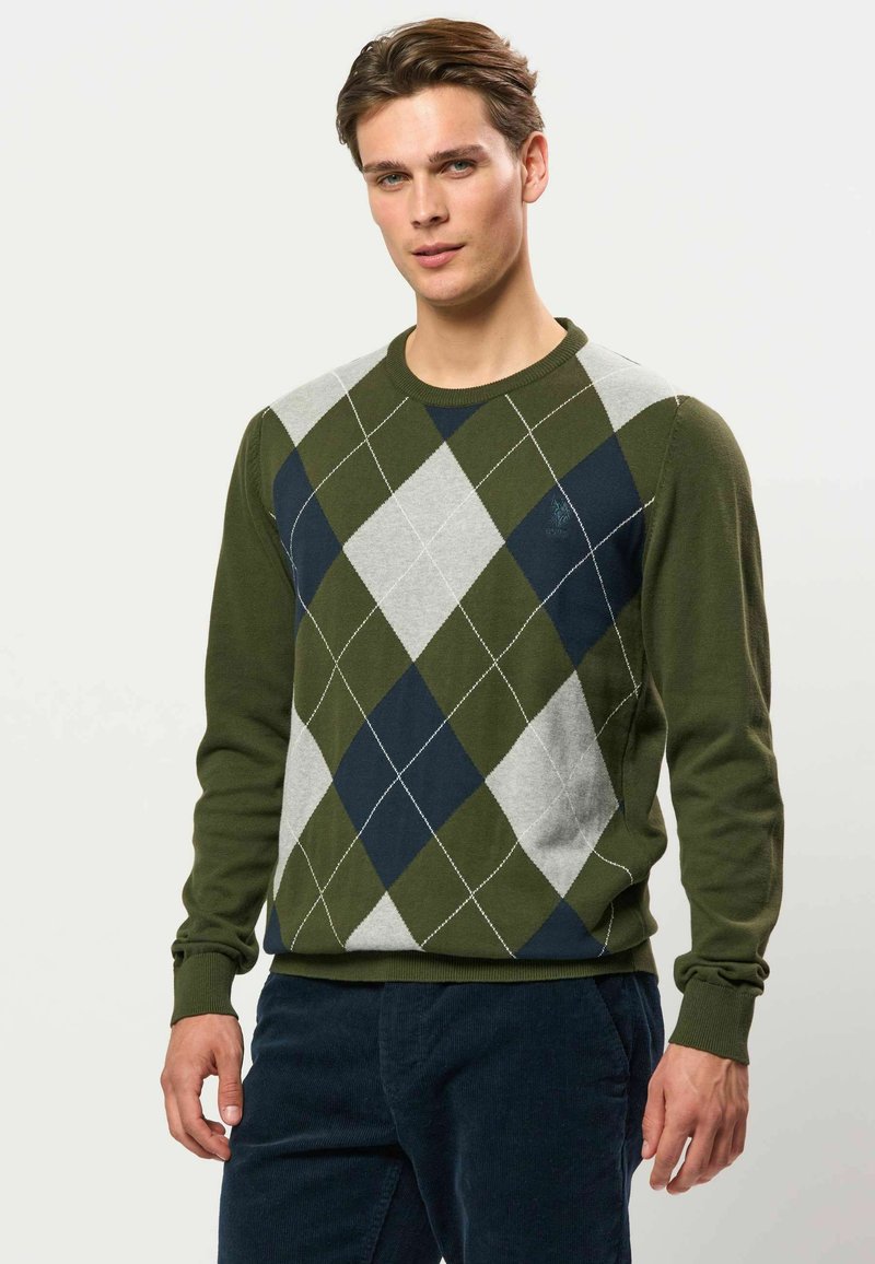 Jeune homme portant un pull à motifs losanges argyle vert, bleu marine et gris et un pantalon sombre, debout devant un fond clair uni.