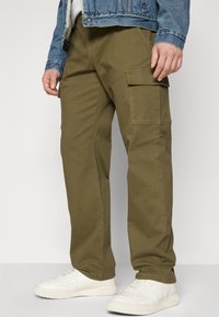 Cargo-Hosen aus olivgrünem Stoff mit geradem Schnitt, ausgestattet mit Seitentaschen, kombiniert mit weißen Sneakers und einer Jeansjacke.