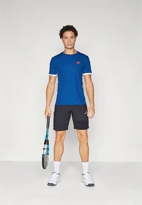 Blå kortermet treningsskjorte med hvite detaljer, kombinert med svarte shorts. Personen holder en tennisracket og har på seg hvite sportssko.