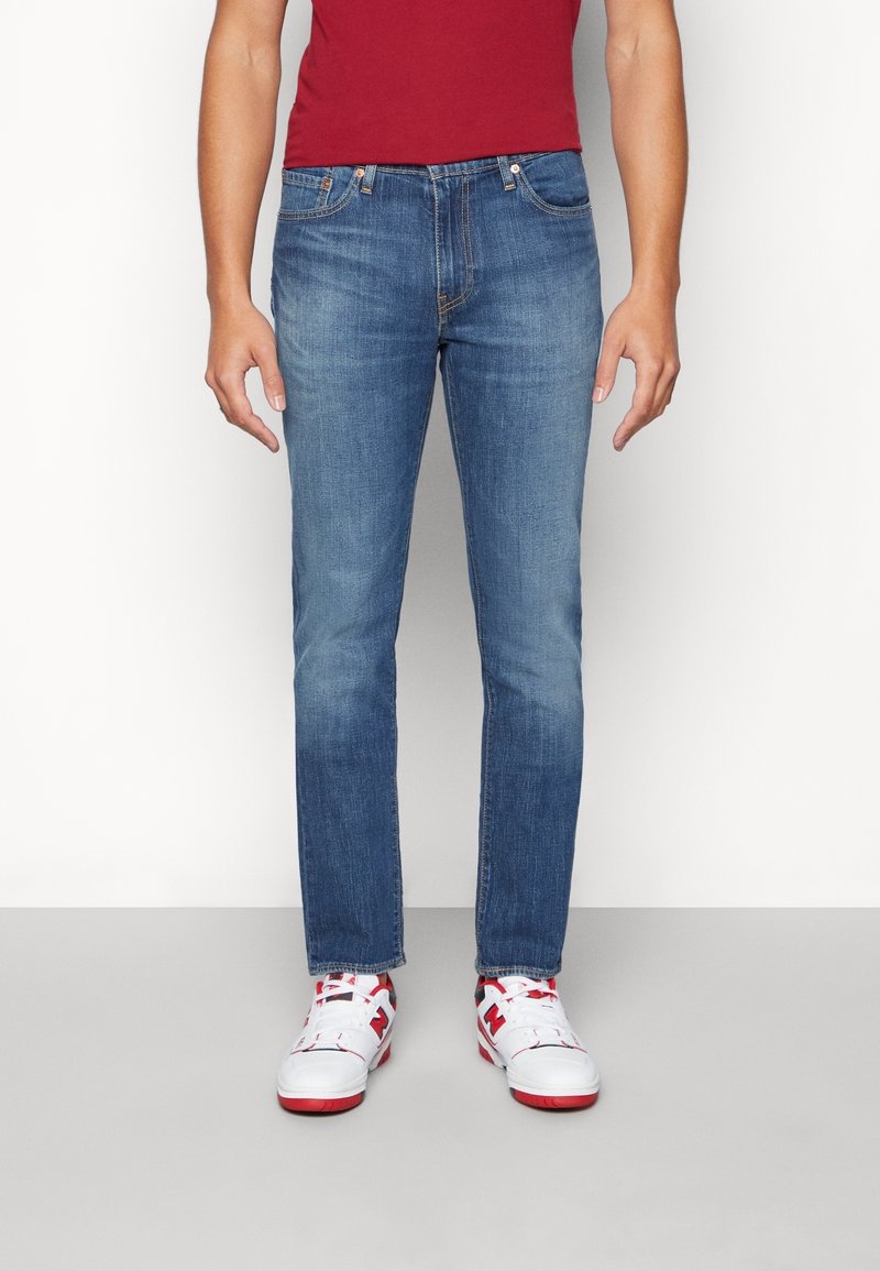 Personne portant un jean slim bleu, une chemise rouge et des baskets blanches avec des accents rouges et noirs, debout devant un fond uni.