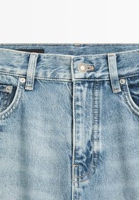 Lichtblauwe spijkerbroek in close-up, met zichtbare voorknop, lussen voor riem en voorzakken met zichtbare stikdetails.