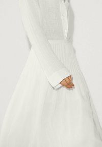 Hvid langærmet skjorte med knaplukning ved kraven og brede manchetter, kombineret med en flydende hvid plisseret nederdel med lodrette linjer.