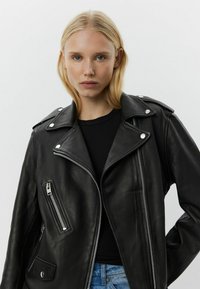 Chaqueta de motorista de cuero negro con herrajes plateados, bolsillos con cremallera en ángulo y solapas anchas. Presenta una textura suave y un diseño ajustado.