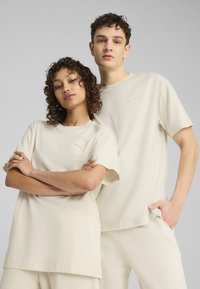 Cremefarvede bomulds T-shirts med korte ærmer. Begge har et diskret broderet logo på brystet. Modellen poserer afslappet med armene krydset.