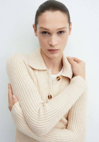 Cardigan a coste ribbed crema con colletto, caratterizzato da tre grandi bottoni marroni, tessuto testurizzato e maniche lunghe.