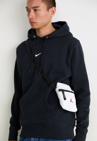 Hvid crossbody taske med en sort lynlås, rødt logo og sort rem. Båret over en mørk hættetrøje med en hvid Nike swoosh. Blå denim jeans.