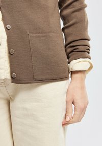 Cardigan marrone a costine con una tasca frontale, con bottoni rotondi e maniche aderenti, indossato sopra una camicia color crema e pantaloni chiari.