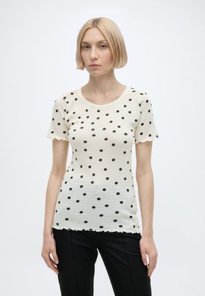 Femme avec une coupe au carré blonde portant un haut blanc à manches courtes avec des pois noirs et un pantalon noir, debout devant un fond uni.