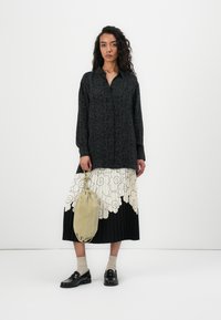 Langärmliges schwarzes Shirt mit floralem Muster, kombiniert mit einem plissierten Schwarz-Weiß-Rock mit großen Blumendesigns. Hält eine beige Tasche.
