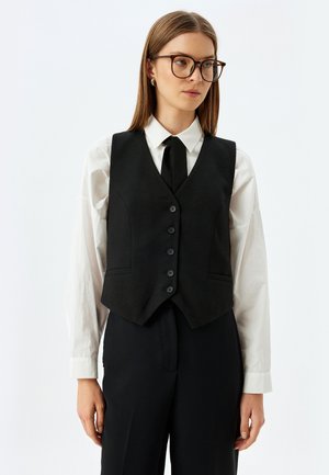 Gilet nero aderente con scollatura a V profonda, cinque bottoni frontali e due tasche laterali, indossato sopra una camicia bianca con una cravatta nera.