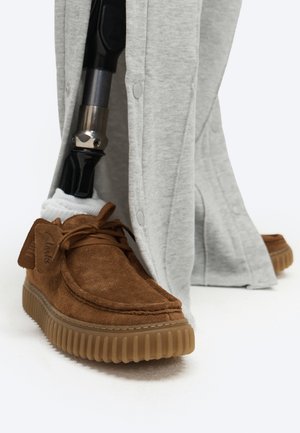 Jambe prothétique avec une chaussette blanche portant une chaussure en daim marron associée à un pantalon gris couvrant la zone de la cheville sur un fond blanc.