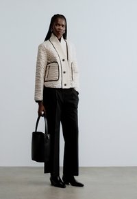 Sieviete valkā krēmkrāsas izšūtu jaku ar melnu apdari, melnas platām kājām bikses, melnus norādītus apavus un tur melnu rokassomu.