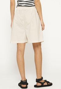 Pantaloni corti beige in cotone con vita elastica, che arrivano a metà coscia, abbinati a sandali neri con cinturini. Tessuto liscio e vestibilità ampia.